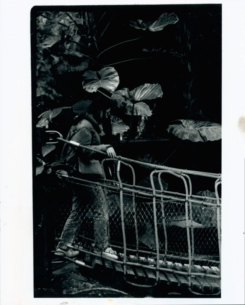 Black and white film print of girl walking across a pedestrian bridge with lush foliage surrounding